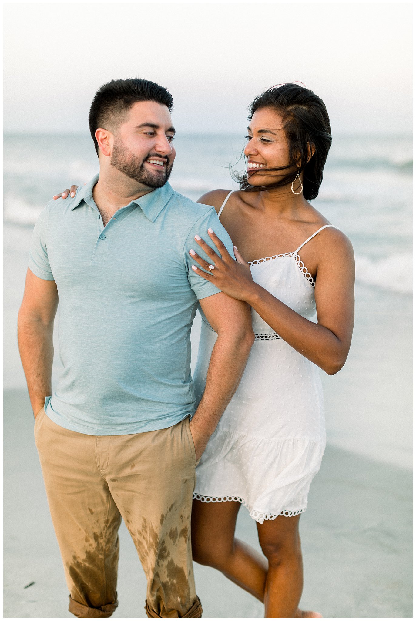 woman in white dress wraps around fiance on the beach and looks at each other with smiles